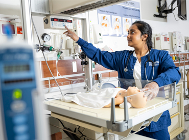 A nursing student reaches across her mannequin infant patient in an incubator, to grab additional medical equipment in the sim lab.